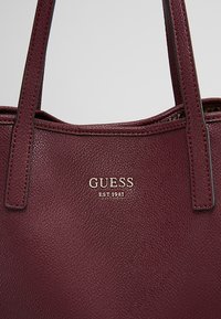 Bordeauxrode leren totebag met een gestructureerde afwerking, voorzien van dubbele handvatten en een goudkleurig geperst "GUESS EST 1981" logo aan de voorkant.