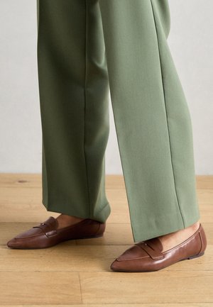 Ruske kožne loafere s oštrim vrhom i suptilnim šavovima, uparene s širokim zelenim hlačama, smještene na drvenom podu.