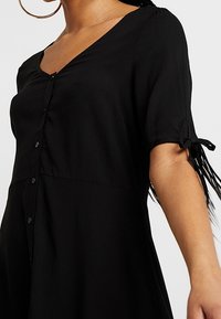 Abito nero con scollatura a V, maniche corte legate ai polsini e dettagli a bottoni verticali. Tessuto liscio con una silhouette fluida.