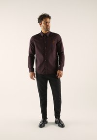 Burgundy long-sleeve shirt with a button-up design, chest pocket, brown buttons, paired with black pants and black shoes. Smooth texture.