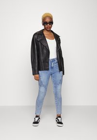 Jaqueta de motociclista de pele preta, t-shirt branca, jeans claros desgastados com detalhes em riscas, ténis pretos com atacadores brancos e óculos de sol.