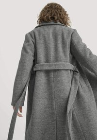 Grauer Wollmischmantel mit langen Ärmeln, tailliertem Gürtel, breitem Kragen und vertikalen Nähten. Der Stoff hat ein weiches, strukturiertes Aussehen.