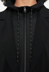 Geacă neagră ușoară, cu guler înalt, fermoar argintiu și șnururi reglabile. Material texturat cu finisaj neted.