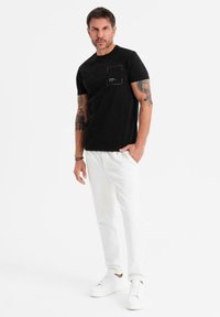 Svart kortärmad t-shirt med bröstficka och subtil textur. Matchad med vita byxor och vita sneakers på en neutral bakgrund.