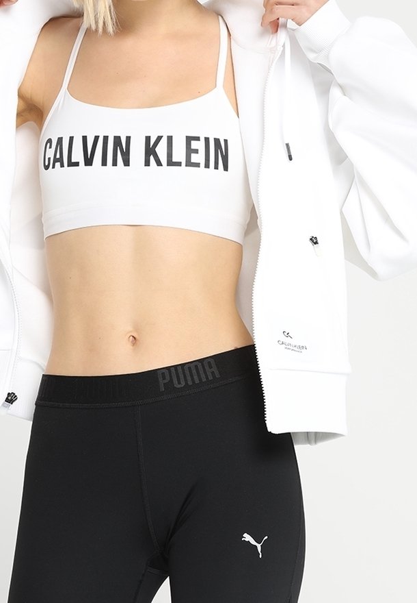 Brassière de sport blanche arborant le logo "CALVIN KLEIN" en noir gras, accompagnée d'un legging noir taille haute avec le logo "PUMA" sur la ceinture.
