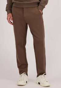 Bruine sportbroek met een textuurafwerking, voorzien van zijzakken en zwarte piping, gecombineerd met witte en zwarte sneakers.