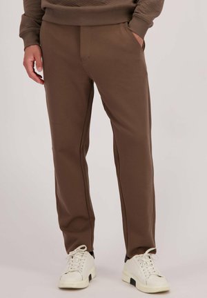 Bruine sportbroek met een textuurafwerking, voorzien van zijzakken en zwarte piping, gecombineerd met witte en zwarte sneakers.