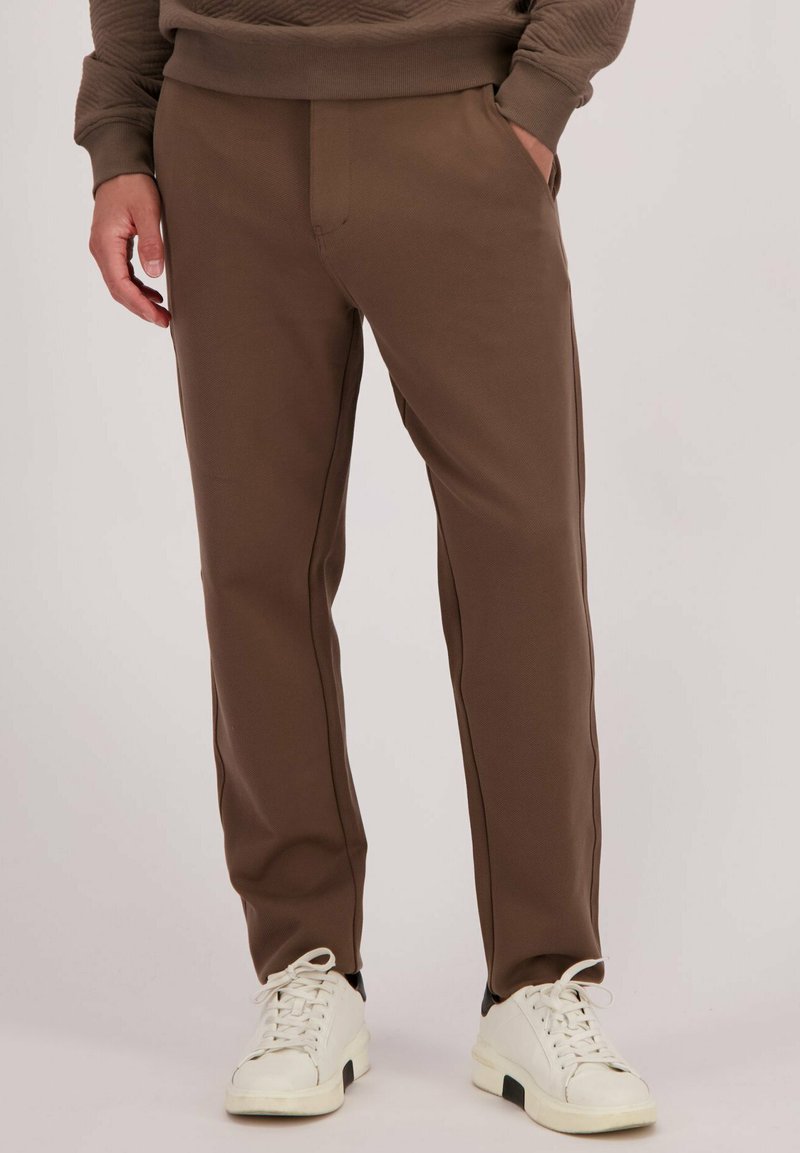 Bruine sportbroek met een textuurafwerking, voorzien van zijzakken en zwarte piping, gecombineerd met witte en zwarte sneakers.