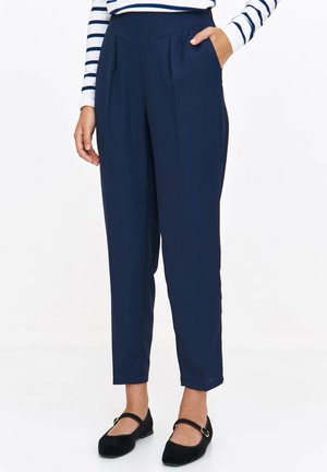 Granatowe spodnie o dopasowanym kroju z plisami z przodu i bocznymi kieszeniami, wykonane z gładkiego materiału. Noszone z czarnymi płaskimi butami.