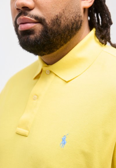 Ανδρας με γένια που φοράει κίτρινο πόλο πουκάμισο με μπλε κεντημένο λογότυπο παίκτη πόλο στο στήθος.