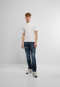 Baltā kokvilnas polo krekla ar trim pogām slēpta plāksne, apvienota ar tumši ziliem nolietotajiem džinsiem un pelēkiem sporta apaviem ar akcentiem.