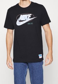 Schwarzes Baumwoll-T-Shirt mit einem großen weißen Nike-Logo und blauen Akzenten sowie dem Text "SINCE 1972". Das Etikett am Saum enthält Größeninformationen.