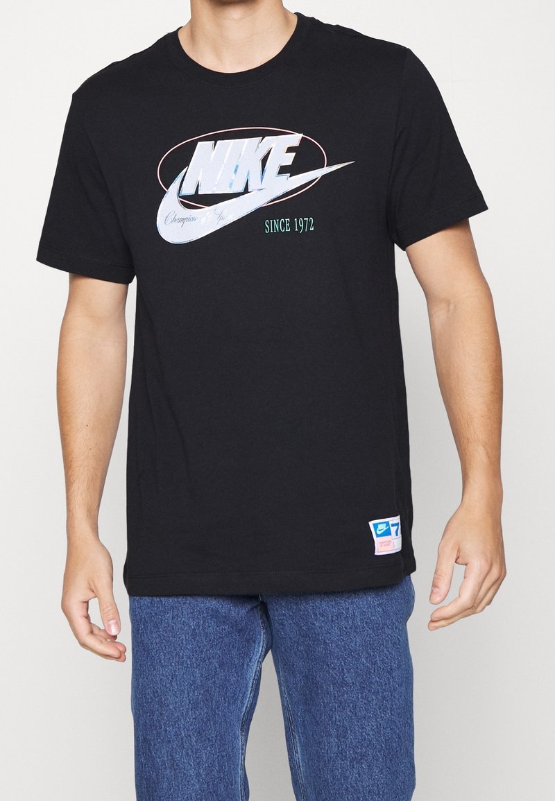 Schwarzes Baumwoll-T-Shirt mit einem großen weißen Nike-Logo und blauen Akzenten sowie dem Text "SINCE 1972". Das Etikett am Saum enthält Größeninformationen.