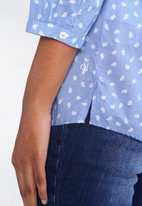 Chemise bleu clair avec motif paisley blanc, manchettes boutonnées et fente sur le côté. Association avec un jean en denim foncé, mettant en valeur un tissu texturé.