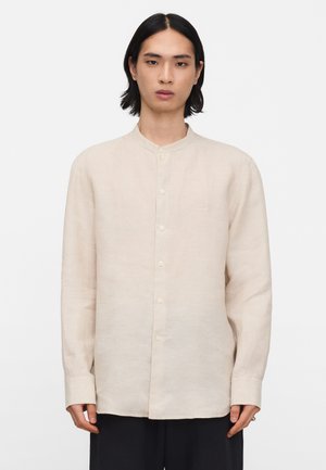 Jonge persoon met steil zwart haar, gekleed in een beige mandarijnkraag overhemd met knopen en lange mouwen en een donkere broek, kijkend naar voren.