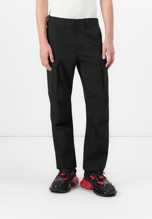 Schwarze Cargo-Hosen aus einem strapazierfähigen Stoff, mit seitlichen Taschen, lockerer Passform und Knopfverschluss. Kombiniert mit schwarz-roten Sneakers.