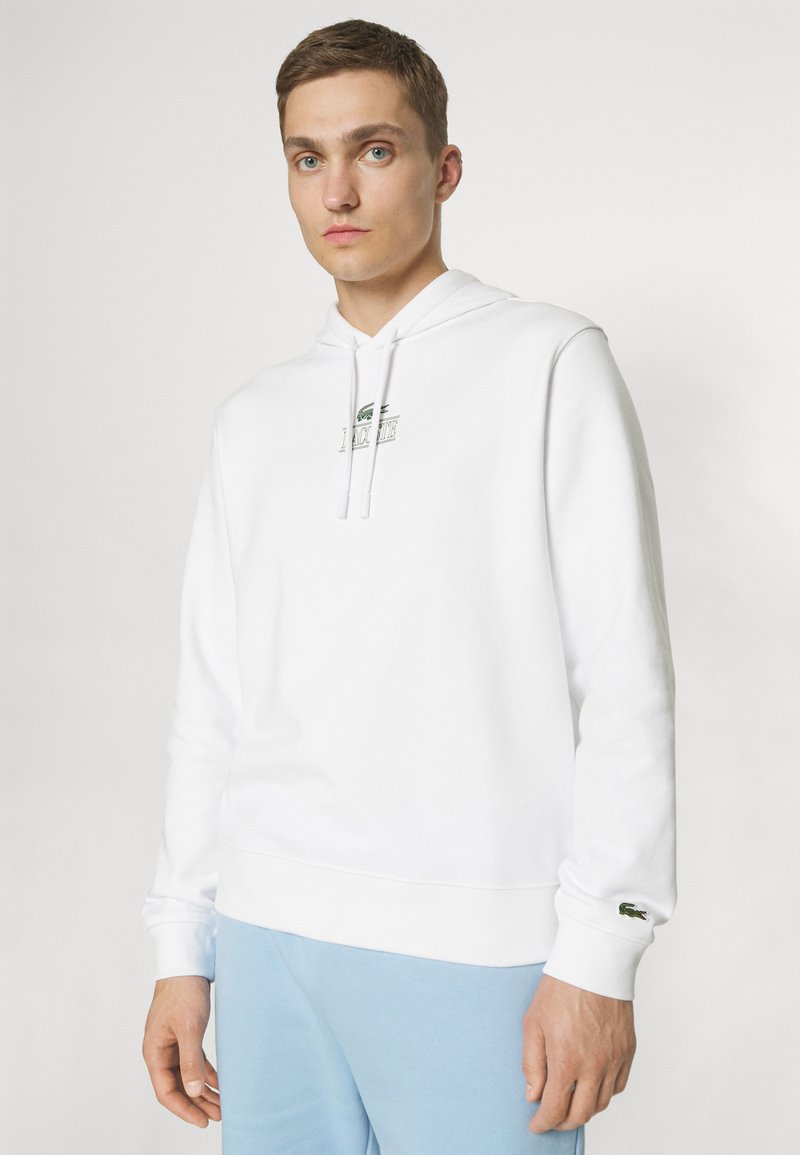 Sweatshirt à capuche blanc avec un logo vert sur la poitrine et un petit logo sur le poignet. Fabriqué en tissu doux et texturé.