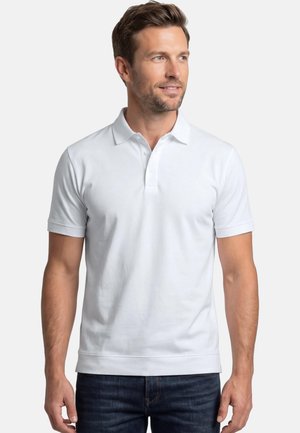 Mężczyzna w gładkiej białej koszulce polo z krótkim rękawem i ciemnych dżinsach, stojący na białym tle, patrzący w prawo.