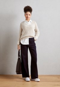 Beżowy sweter z prążkowanej dzianiny, biała koszula z guzikami, szerokie ciemne spodnie, białe sneakersy i brązowa torba. Tło jest jasnoszare z drewnianą podłogą.