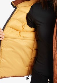 Personne portant un haut noir tenant une veste marron ouverte montrant son intérieur matelassé marron clair et sa poche intérieure.