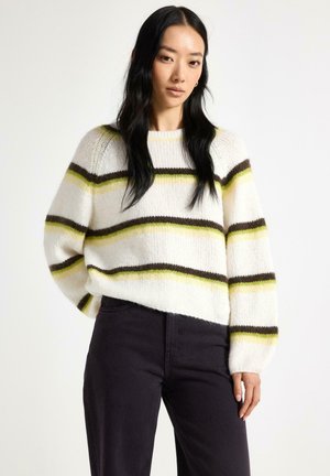 Kvinde med langt sort hår iført en hvid strikket sweater med grønne og brune striber og sorte bukser, stående mod en ensfarvet baggrund.