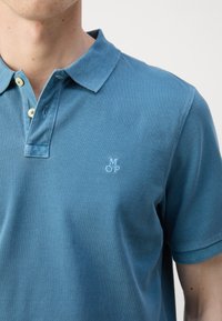 Homme portant un polo bleu à manches courtes avec deux boutons beige et un petit logo brodé blanc "M O P" sur la poitrine.