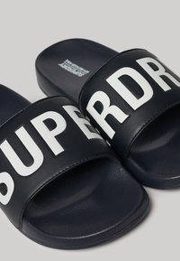 Par par navy blue slide sandaler med brede sorte stropper, der har stort hvidt "SUPERDRY" tekst henover begge stropper.