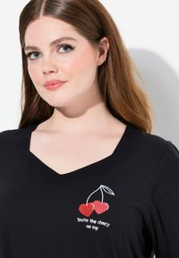 Maglietta nera a V con ciliegie rosse ricamate e la scritta "Sei la ciliegina sulla torta" in bianco. Tessuto morbido, vestibilità rilassata, design casual.