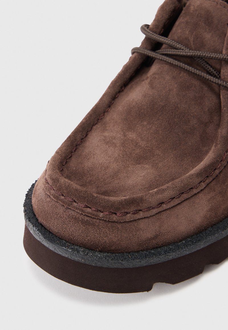 Bruine suède laars met een ronde neus, contrasterende stiksels en een dikke zwarte rubberen zool. Veters zijn bruin en door oogjes gestrikt.