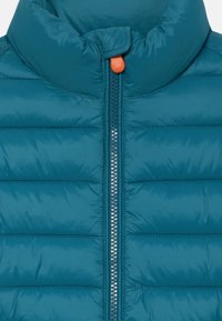 Vesta puf cu culoare teal, cu un guler înalt, design cu cusături, și un fermoar argintiu. Are un tab de tragere portocalie și o textură netedă, lucioasă.