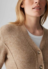 Cardigan beige lavorato a maglia con scollo a V, caratterizzato da una finitura testurizzata e tre grandi bottoni. Camicia interna bianca a coste visibile sotto.