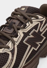 Temno rjav športni copat znamke New Balance z mrežasto tkanino, kremnimi plastmi, rjavimi vezalkami in logotipom na strani ter jeziku.