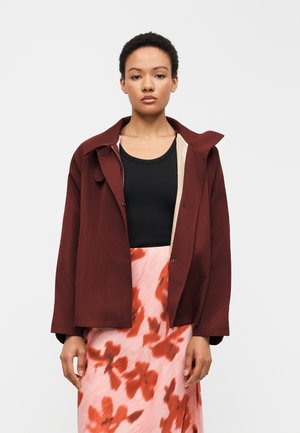 Γυναίκα που φοράει σκούρο κόκκινο ανοιχτό σακάκι πάνω από μαύρο τοπ και ροζ φούστα με κόκκινα λουλουδάτα σχέδια, στέκεται μπροστά σε απλό φωτεινό φόντο.