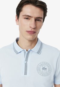 Ljusblå pikétröja med ribbat krage som har svarta ränder, knapplist och broderad Lacoste-logotyp på bröstet.