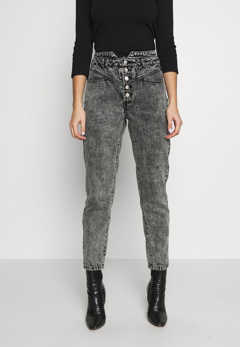 Femme portant un jean gris taille haute effet délavé avec braguette à boutons et des bottines noires, debout sur un fond clair uni.