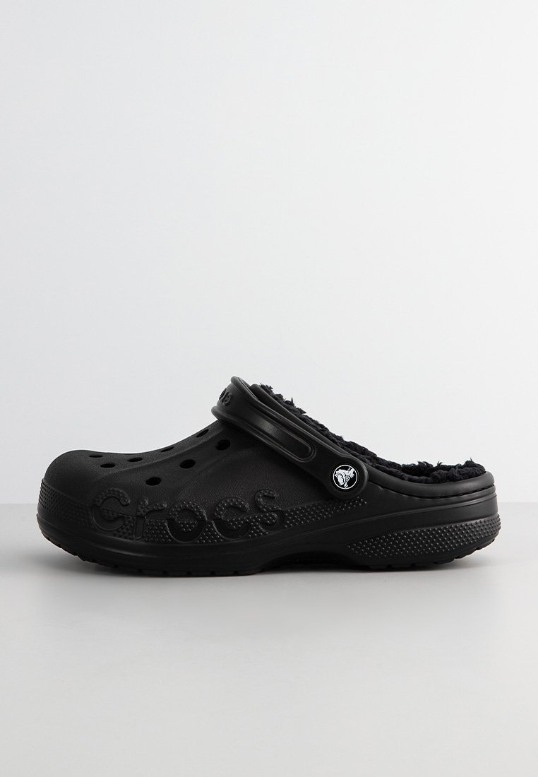 Zueco negro Crocs con forro de felpa, agujeros de ventilación y correa ajustable en el talón, mostrado de lado sobre una superficie lisa.