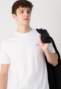 Weißes T-Shirt mit Rundhalsausschnitt, das ein kleines rotes Logo-Patch trägt. Die Person hält ein gefaltetes schwarzes Kleidungsstück über der Schulter.