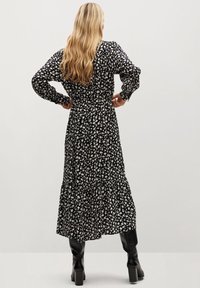 Schwarzes Blumenkleid mit langen Ärmeln, tailliertem Schnitt und schichtenförmigem Rock. Das Material hat eine weiche Textur und ein kleines weißes Blumenmuster. Schwarze knielange Stiefel.