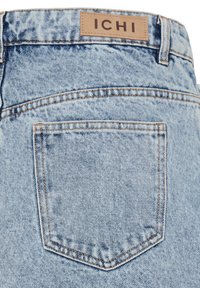 Poche arrière de jean en denim bleu clair avec surpiqûres contrastées et étiquette de marque en cuir marron indiquant "ICHI" sur la ceinture.