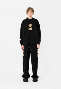 Conjunto de sudadera y pantalones negros con un diseño de oso de peluche en una tela texturizada. La sudadera tiene mangas largas, mientras que los pantalones tienen detalles en los bolsillos.