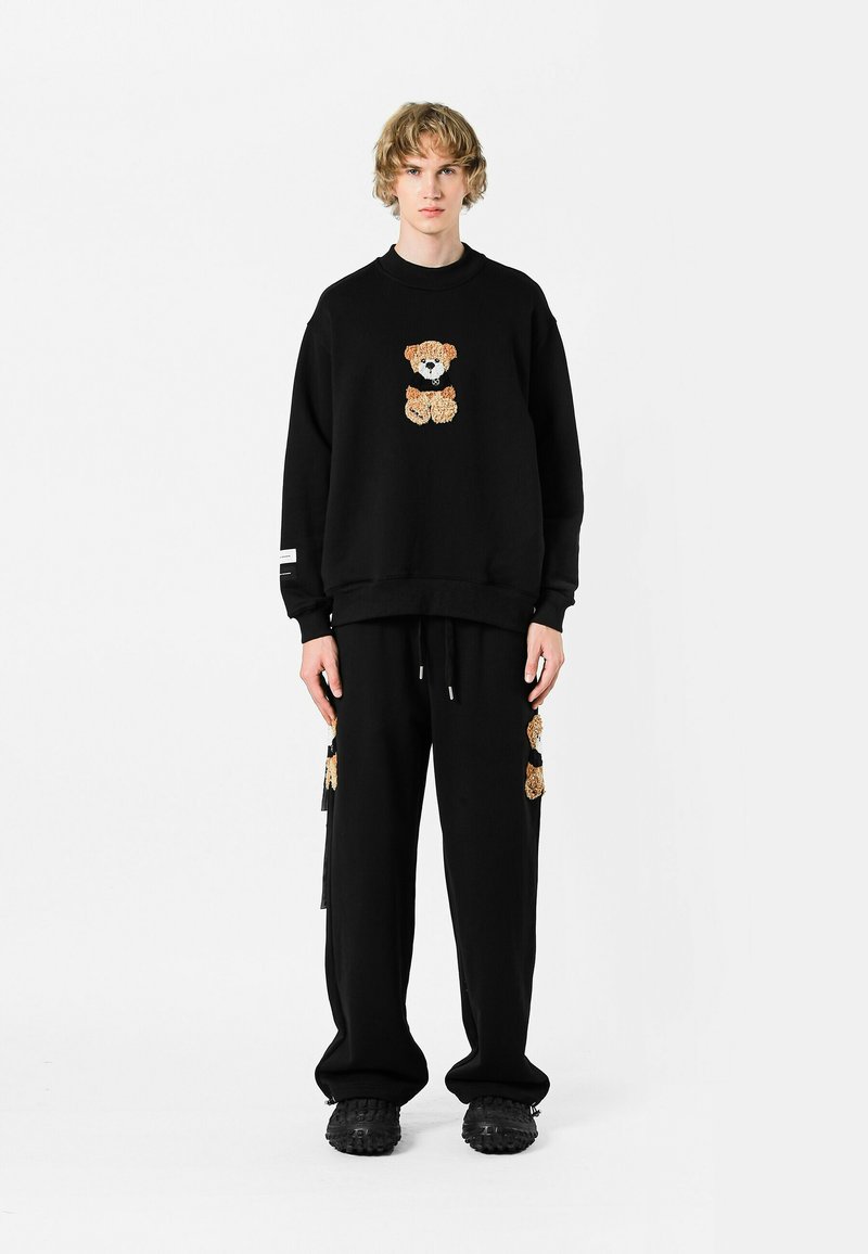 Conjunto de sudadera y pantalones negros con un diseño de oso de peluche en una tela texturizada. La sudadera tiene mangas largas, mientras que los pantalones tienen detalles en los bolsillos.