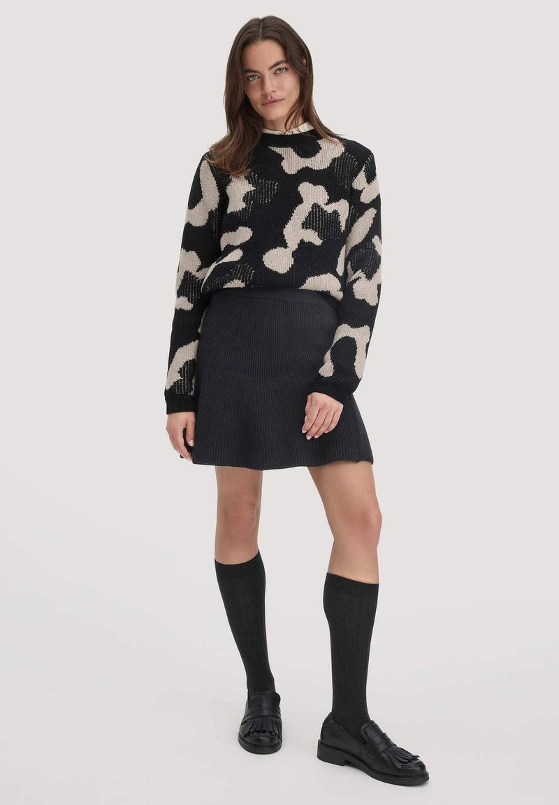 Zwart en beige gebreide trui met abstract patroon, gecombineerd met een zwarte geribbelde rok, kniehoge zwarte sokken en zwarte instappers.