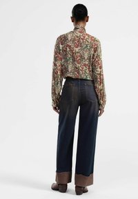 Blusa beige con motivo floreale e colletto alto, abbinata a pantaloni a gamba larga in denim scuro con vita alta e orli consumati di colore marrone.