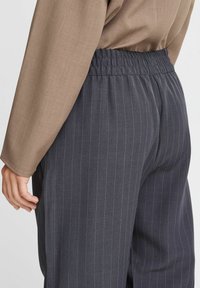 Gray pinstriped trousers with an elastic waistband, paired with a light brown, textured long-sleeve top, shown from the side.