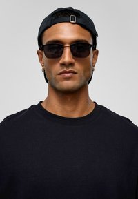 Zwarte zonnebril met rechthoekige lenzen, een zwarte pet met een gesp, en een zwart T-shirt met ronde hals. Glad textuur en minimalistisch ontwerp.