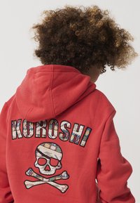 Otrok s kodrastimi lasmi nosi rdečo kapuco z motivom lobanje in prečkami ter z vzorčastim vezenjem besedila "KUROSHI" na hrbtni strani.
