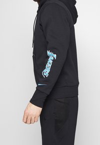 Sudadera negra con gráfico azul en la manga izquierda, que cuenta con un bolsillo frontal y cordones ajustables. Tela texturizada y diseño ajustado.