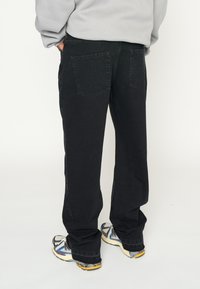 Pantalones de mezclilla negros con corte recto, que cuentan con dos bolsillos traseros y un acabado ligeramente texturizado. Llevados con zapatillas beige y azules.