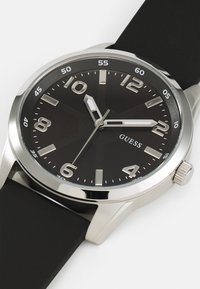 Montre à cadran noir avec index et aiguilles argentés. Boîtier en acier inoxydable, bracelet en caoutchouc noir, fond texturé sur le cadran.