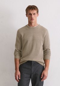 Pull beige texturé avec un col rond et des poignets côtelés, associé à un pantalon gris foncé. Détail logo subtil sur la poitrine.